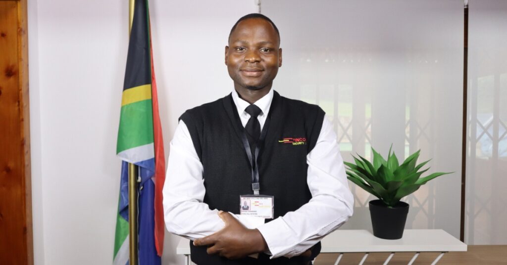 A professional male security officer from Tincol 24/7 Security stands confidently in uniform with arms folded, wearing an ID badge, in front of a South African flag and office background with indoor plants.