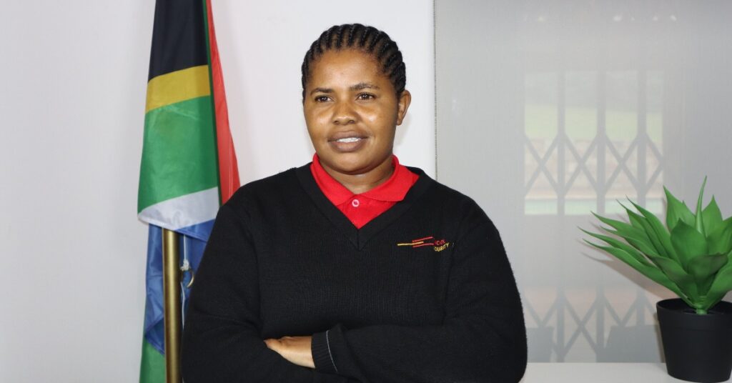 A female Tincol 24/7 Security officer poses confidently indoors in front of a South African flag and a potted plant, wearing the company’s branded black and red uniform.