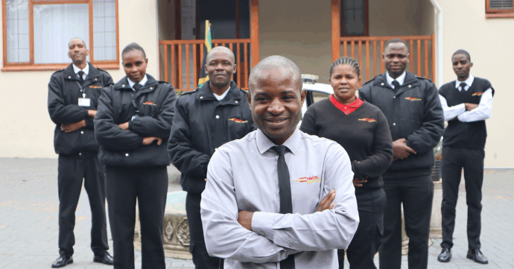 A group of uniformed security personnel standing together outside a building, with one team member in front smiling confidently while the rest stand behind with folded arms, representing a united security team.