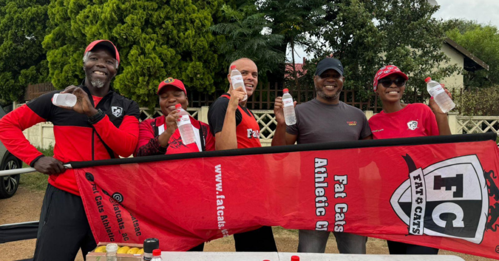 A group of five smiling people wearing red and black sportswear stand outdoors holding water bottles behind a red banner for Fat Cats Athletic Club.