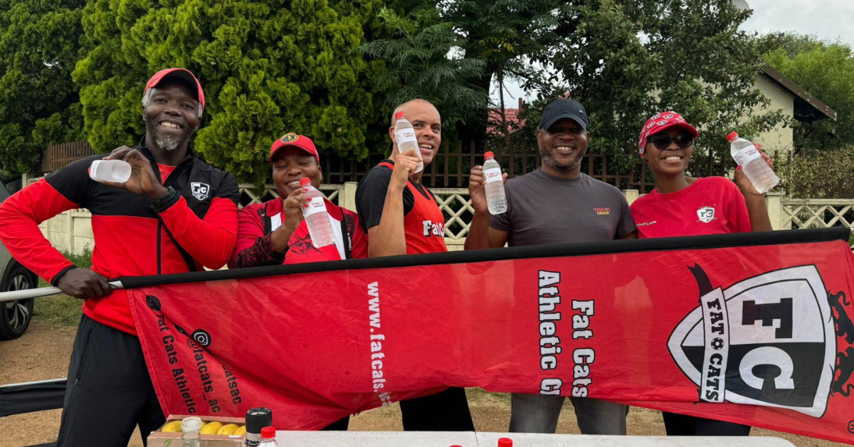 A group of five smiling people wearing red and black sportswear stand outdoors holding water bottles behind a red banner for Fat Cats Athletic Club.