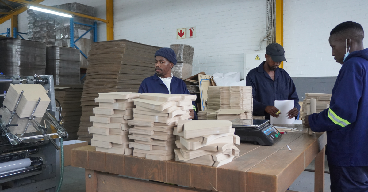 Three factory workers in blue uniforms sorting and stacking beige paper packaging materials on a wooden table in an industrial workspace, with machinery and large stacks of cardboard visible in the background.
