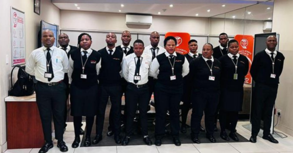 A group of uniformed security staff stand in a line indoors, wearing black vests, white shirts, black ties, and ID badges. They face the camera in a well-lit office or reception area with branded signage in the background.