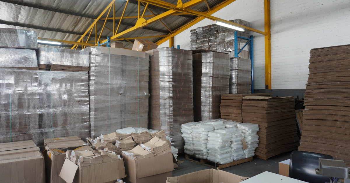 Warehouse interior with stacked manufactured consumables, including bundled geo-sample envelopes, inner and outer boxes and plastic materials, arranged for storage and distribution.