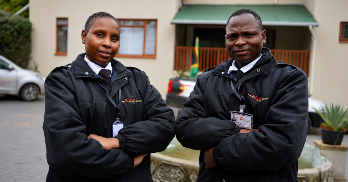 Two uniformed security officers standing outdoors with their arms crossed in front of a residential-style building. Both are wearing black branded jackets, ties, and ID badges, presenting a professional and confident appearance.